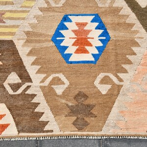 May include: A rug with a geometric pattern in shades of brown, blue, red, and white. The rug has a fringed edge and is made of a woven fabric.