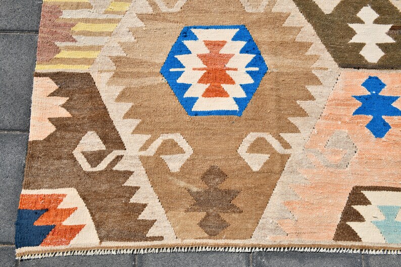 May include: A rug with a geometric pattern in shades of brown, blue, red, and white. The rug has a fringed edge and is made of a woven fabric.