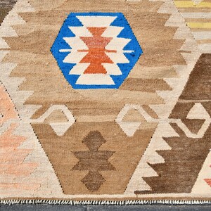 May include: A close-up of a woven rug with a geometric pattern in shades of brown, beige, white, blue, orange, and red. The rug has a fringe border.