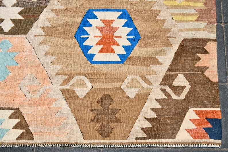 May include: A close-up of a woven rug with a geometric pattern in shades of brown, beige, white, blue, orange, and red. The rug has a fringe border.