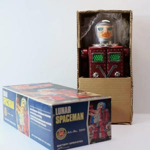Pode incluir: Um brinquedo de robô Lunar Spaceman vermelho vintage em sua caixa de papelão original. O robô tem uma cabeça de cúpula transparente, luzes verdes e um rosto prateado. A caixa é azul e apresenta as palavras "Lunar Spaceman" e ilustrações do robô.