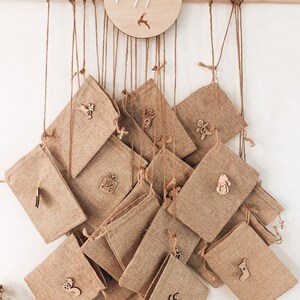 May include: A burlap advent calendar with small, numbered pouches hanging from twine. A wooden circle at the top displays the name "Pippa" and a reindeer. Each pouch has a wooden Christmas-themed embellishment.
