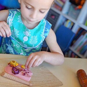 May include: A child is decorating pink play dough with dried flowers. The flowers are yellow, pink, and red. The child is wearing a blue and green floral dress.