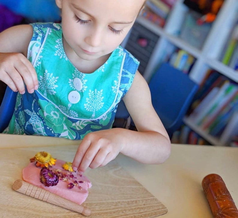 May include: A child is decorating pink play dough with dried flowers. The flowers are yellow, pink, and red. The child is wearing a blue and green floral dress.