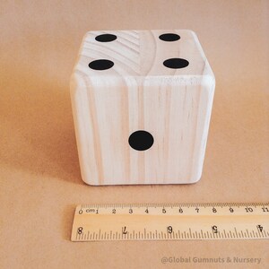 May include: A large wooden die with black dots, showing the numbers one and four. The die is light-colored wood and is approximately 10 cm in size. A wooden ruler is placed next to the die for scale.