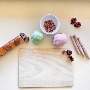 May include: A wooden rolling pin with a leaf design, a small wooden rolling pin, a wooden chopping board, three wooden dowels, two red ladybug ornaments, a bowl of dried rose petals, and two balls of play dough, one green and one pink.