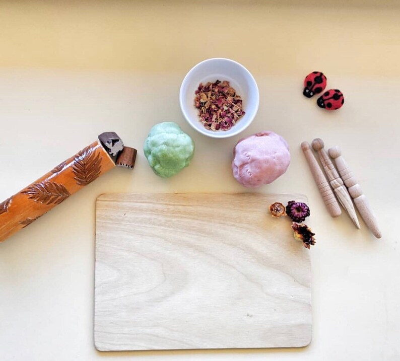 May include: A wooden rolling pin with a leaf design, a small wooden rolling pin, a wooden chopping board, three wooden dowels, two red ladybug ornaments, a bowl of dried rose petals, and two balls of play dough, one green and one pink.
