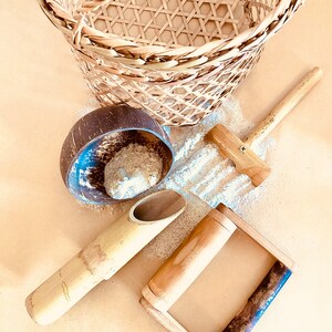 May include: A woven basket, coconut shell bowl, wooden mallet, bamboo scoop, and a wooden frame with a handle are arranged on a light brown surface. The items are scattered with a light-coloured substance, possibly sand or flour. The items are natural in colour.