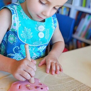 May include: A child wearing a blue and turquoise floral dress uses a wooden tool to press into pink play dough with dried flower petals on top.