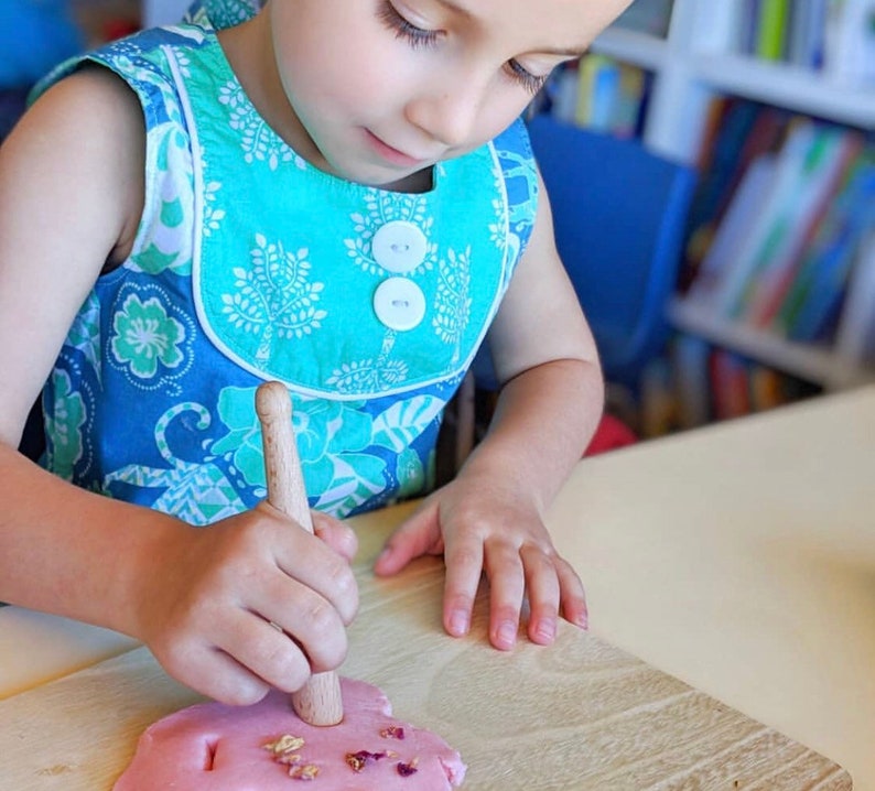 May include: A child wearing a blue and turquoise floral dress uses a wooden tool to press into pink play dough with dried flower petals on top.
