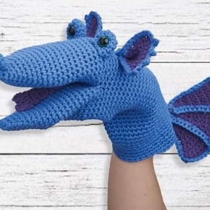 May include: Blue crocheted puppet dragon with purple wings and green eyes. The dragon has a large mouth and is ready to be used for puppet shows.