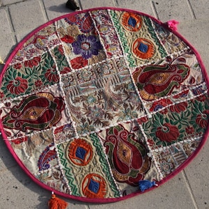 Op de afbeelding: Een ronde patchwork kussen met een roze rand en kleurrijke kwastjes. Het kussen heeft geborduurde stoffen vierkanten in rood, groen, blauw en crème, met bloemen- en paisley patronen. Ongeveer 70 cm in diameter.