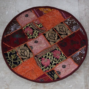Op de afbeelding: Een ronde patchwork vloerkussen met een bordeauxrode rand. Het kussen heeft een verscheidenheid aan stoffen in tinten oranje, roze, bordeauxrood en goud, met bloemen en geometrische patronen. Het kussen heeft een diameter van ongeveer 61 cm.