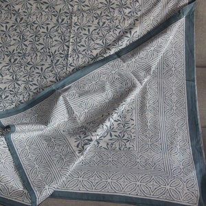 Peut inclure: Nappe pliée, blanc cassé, ornée d'un motif floral bleu-gris. La bordure présente un motif géométrique de la même couleur. Les bords sont finis par une bordure bleu-gris uni.