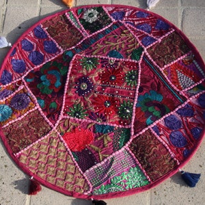Op de afbeelding: Een ronde patchwork vloerkussen met een levendige mix van kleuren. Het kussen heeft een roze rand en is versierd met verschillende geborduurde stoffen patches in roze, paarse, groene en gouden tinten. Decoratieve kwastjes zijn rond de rand bevestigd.