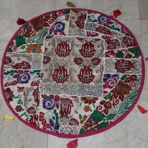 Op de afbeelding: Rond vloerkussen met een patchwork ontwerp. Het kussen heeft een centraal vierkant met een bloemenpatroon in rood en beige, omringd door verschillende stoffen patches met bloemen- en paisley motieven. Het heeft een roze rand en kleurrijke kwasten.