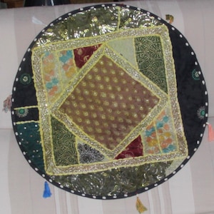 Peut inclure: Un coussin rond et décoratif avec un motif patchwork. Le coussin présente un carré central avec un tissu marron à motifs, entouré d'un carré de patchwork coloré. Le bord extérieur est noir avec des points blancs et de petits pompons.