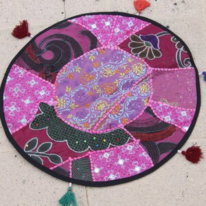 Op de afbeelding: Een ronde patchwork vloerkussen in roze, paarse en bordeaux tinten. Het kussen heeft een verscheidenheid aan stoffen met patronen, waaronder bloemen- en paisley-ontwerpen. Het heeft een zwarte rand en is versierd met kleurrijke kwastjes.