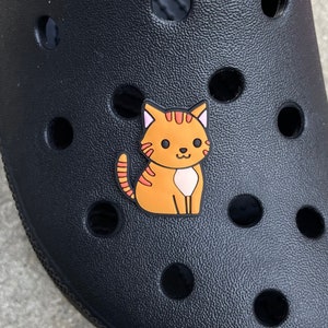 May include: A black shoe with a textured surface and round holes. A small, orange cat charm with black stripes and a white belly is attached to the shoe. The cat charm is a shoe accessory.