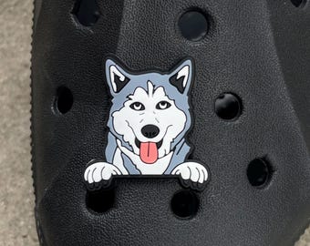 May include: A black shoe with a decorative charm featuring a cartoon husky dog. The charm is in shades of gray, white, and pink, with the dog's tongue sticking out. The shoe has multiple holes.