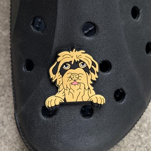 May include: A black clog with a decorative dog charm. The charm is a cartoon-style dog with tan fur, black ears, and a pink tongue. The clog has multiple ventilation holes.