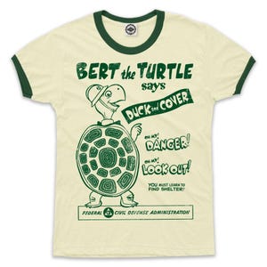 May include: A vintage-style t-shirt with a green ringer neck. The shirt features a cartoon turtle wearing a helmet and holding a sign that says "BERT the TURTLE says DUCK and COVER". The sign also includes the text "OH MY! DANGER", "OH MY! LOOK OUT!" and "YOU MUST LEARN TO FIND SHELTER!" The bottom of the sign reads "FEDERAL CIVIL DEFENSE ADMINISTRATION".