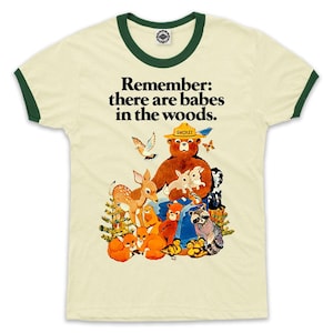 May include: A white t-shirt with a green ringer, featuring a cartoon illustration of Smokey Bear with a group of animals. The text "Remember: there are babes in the woods." is printed on the shirt.