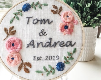 May include: Embroidered hoop with the names "Tom & Andrea" and the date "Est. 2019" in gray thread. The hoop is decorated with pink and blue flowers and brown and green leaves.