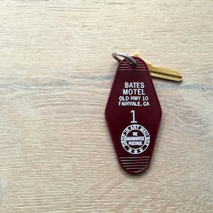 May include: A red motel key with a gold key ring and a key. The key has the text "Bates Motel, Old Hwy 10, Fairvale, CA" and the number "1". The key also has a circle with the text "Drop In Any Mailbox, We Guarantee Postage".