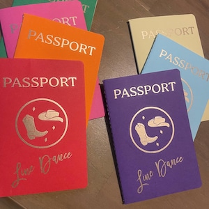 May include: A collection of colorful passport-sized notebooks. The notebooks are in various colors, including red, purple, orange, pink, green, and blue. Each notebook has the word "PASSPORT" printed on the cover, along with a graphic and the words "Line Dance".