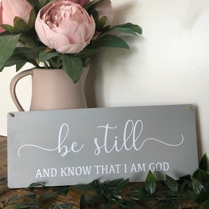 May include: A rectangular, grey wooden sign with the words "be still" and "and know that I am God" in white script. The sign is decorated with a vase of pink flowers and greenery.