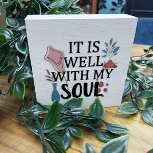 May include: A white wooden block featuring the phrase "IT IS WELL WITH MY SOUL" in black lettering. The block is adorned with floral and vase illustrations in soft hues. Green foliage surrounds the block, set on a wooden surface.