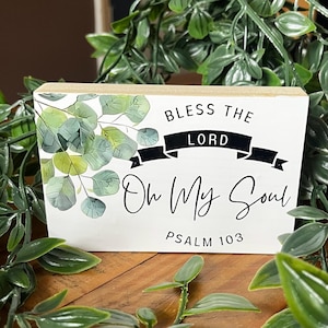 May include: A rectangular wooden sign with the text "BLESS THE LORD Oh My Soul PSALM 103". The sign has a white background with black lettering and a banner. It features a watercolor eucalyptus design in green and grey.