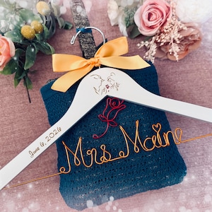 May include: A white wooden wedding dress hanger with the text "June 6, 2026" and "Mrs. Maine" in gold wire. The hanger has a yellow bow and a design of a couple and a rose. A blue sweater is on the hanger.
