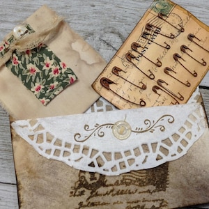 May include: An assortment of vintage-style paper ephemera. Includes a small fabric pouch with a floral tag, a postcard with safety pins, and a decorative envelope with a lace overlay and wax seal.