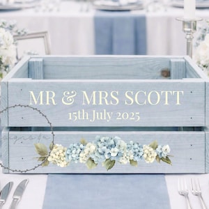 May include: A pale blue wooden crate, inscribed with "MR & MRS SCOTT 15th July 2025" in gold. It features a floral design of blue and white hydrangeas. The crate is part of a wedding table setting.