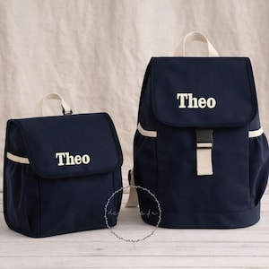 May include: Two navy blue backpacks with white detailing and the name "Theo" embroidered on the front. The larger backpack has a flap closure with a buckle, the smaller one has a flap closure.