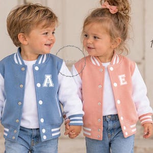 May include: A blue varsity jacket with white sleeves and a white letter A, and a peach varsity jacket with white sleeves and a white letter E. Both jackets feature white snap buttons, striped ribbed cuffs, and the text Little Adventures Big Memories.