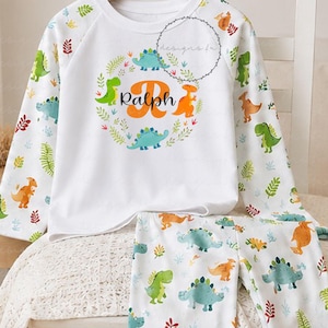 May include: A white long-sleeve shirt and matching pants set featuring a colorful dinosaur and leaf pattern. The shirt displays a central graphic with a large orange letter R, the name Ralph in black script, and a circular wreath of dinosaurs.
