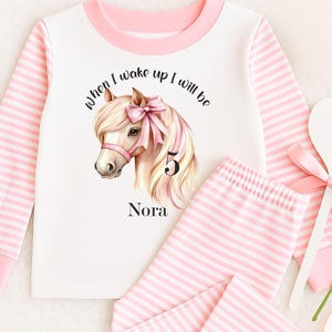 May include: White and pink striped pajama set. The top features a horse illustration with a pink bow and the text "When I wake up I will be 5 Nora". The set includes a long-sleeved top and matching pants.