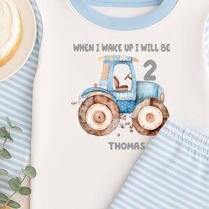 May include: White and light blue pajama set with a tractor graphic. The top has a blue collar and sleeves with the text "WHEN I WAKE UP I WILL BE 2 THOMAS". A birthday cake with blue candles and a star wand are also in the image.