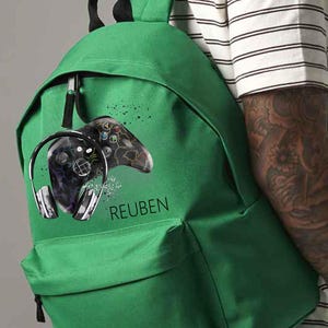 May include: Green backpack with a gaming controller and headphones design. The backpack has the name "REUBEN" printed on it. The design is in black and grey, with a splatter effect. The backpack is made of a durable material and has a front pocket.