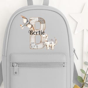 May include: A pale grey backpack with a front zip pocket and adjustable straps. The backpack has a decorative letter "B" with a dinosaur and airplane design, and the name "Bertie". A star-shaped wand and a sprig of greenery with a gold bow are next to the backpack.