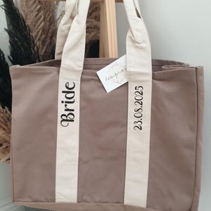 May include: A beige canvas tote bag with cream-coloured straps. The word "Bride" is printed vertically on one strap, and "23.08.2025" is printed on the other. A small tag hangs from the strap. The bag is designed for a wedding.
