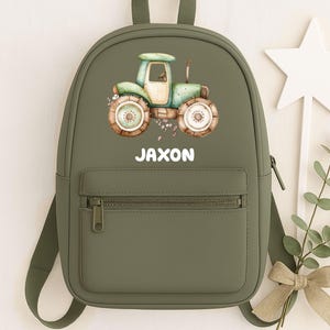 May include: An olive green backpack with a tractor illustration and the name "JAXON". The tractor is in shades of green and brown. The backpack has a front pocket and adjustable straps. A star wand and sprig of greenery are next to the backpack.