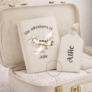 May include: An open cream suitcase displays a passport cover and luggage tag. The passport cover has an airplane design and the text "The adventures of Alfie." The luggage tag is labelled "Alfie."