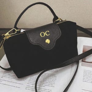 May include: Black handbag with a top handle and a detachable shoulder strap. The bag has a flap closure with gold-coloured hardware and the initials "OC". The bag is on top of printed paper.