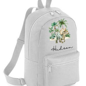 May include: A grey canvas backpack with a dinosaur and palm trees design. The backpack has a zipped pocket on the front and the name "Hudson" is printed on the bottom.
