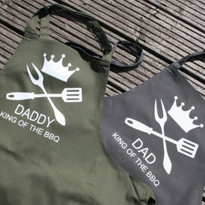 May include: Two green and black aprons with white text and graphics. The aprons say "Daddy King of the BBQ" and "Dad King of the BBQ".