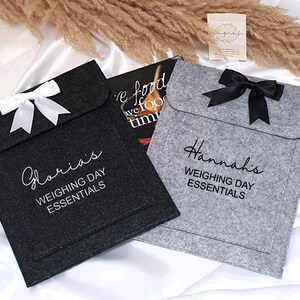 May include: Two gray felt pouches with black ribbon bows. The pouches are personalized with the names "Gloria's" and "Hannah's" and the text "Weighing Day Essentials".